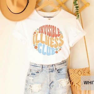 K&ouml;nnte beinhalten: Wei&szlig;es T-Shirt mit einer bunten Grafik, auf der "Invisible Illness Club" mit Blumen um den Text steht.