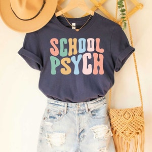 May include: A navy blue t-shirt with the text "SCHOOL PSYCH" in rainbow-colored letters. The shirt is worn with denim shorts and a woven bag.
