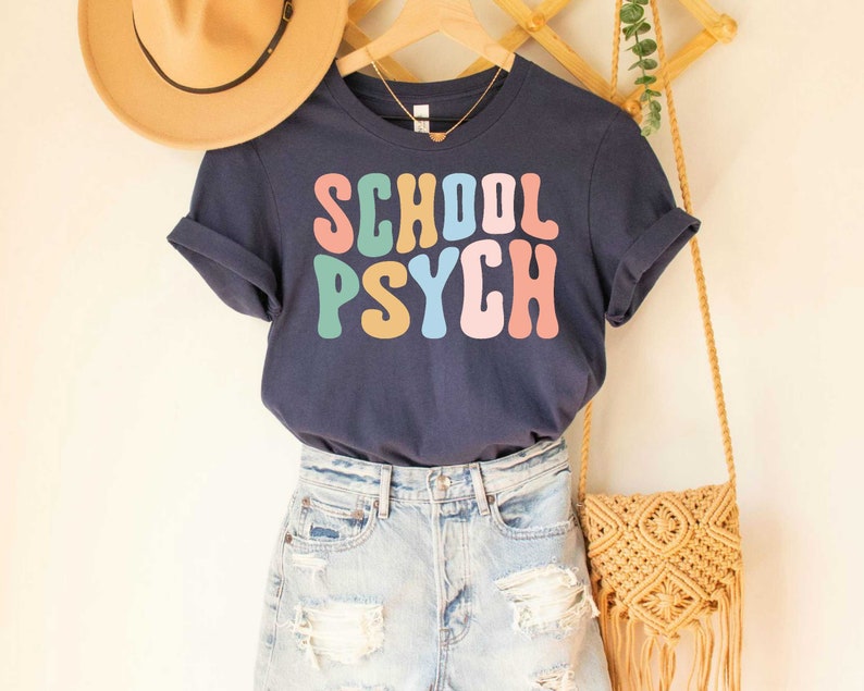 May include: A navy blue t-shirt with the text "SCHOOL PSYCH" in rainbow-colored letters. The shirt is worn with denim shorts and a woven bag.