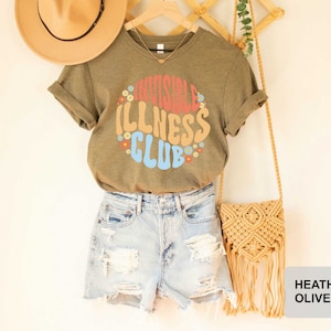 K&ouml;nnte beinhalten: Ein heidefarbenes olivgr&uuml;nes T-Shirt mit dem Text "Invisible Illness Club" in einer Retro-Schriftart mit bunten Blumen, die den Text umgeben. Das Shirt wird von einer Person mit Jeans-Shorts und einem Strohhut getragen.