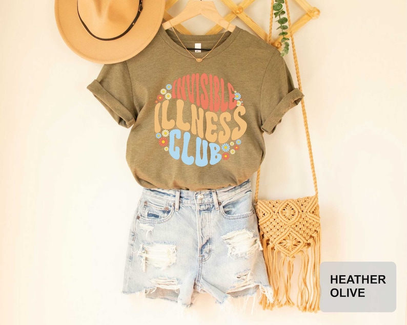 K&ouml;nnte beinhalten: Ein heidefarbenes olivgr&uuml;nes T-Shirt mit dem Text "Invisible Illness Club" in einer Retro-Schriftart mit bunten Blumen, die den Text umgeben. Das Shirt wird von einer Person mit Jeans-Shorts und einem Strohhut getragen.
