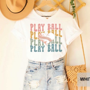 Peut inclure: T-shirt blanc avec un graphique r&eacute;tro d'un ballon de baseball en forme de c&oelig;ur et le texte "PLAY BALL" r&eacute;p&eacute;t&eacute; dans diff&eacute;rentes couleurs.