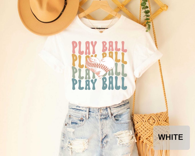 Peut inclure: T-shirt blanc avec un graphique r&eacute;tro d'un ballon de baseball en forme de c&oelig;ur et le texte "PLAY BALL" r&eacute;p&eacute;t&eacute; dans diff&eacute;rentes couleurs.