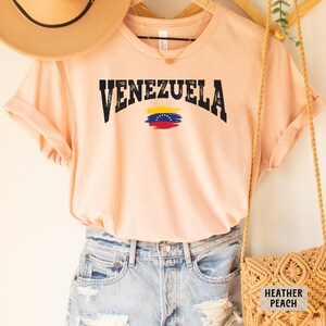 May include: A peach colored t-shirt with the word "VENEZUELA" in black letters with a distressed look. The Venezuelan flag is printed in the center of the shirt.