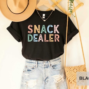 Puede incluir: Camiseta negra con el texto "SNACK DEALER" en colores arco&iacute;ris. La camiseta est&aacute; combinada con un sombrero de paja, pantalones cortos de mezclilla y una bolsa tejida.
