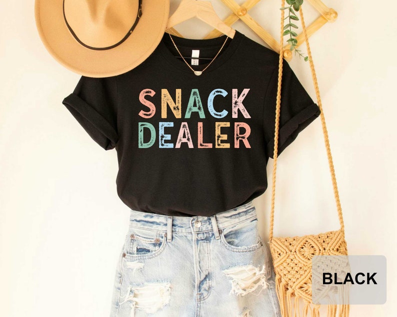 Puede incluir: Camiseta negra con el texto "SNACK DEALER" en colores arco&iacute;ris. La camiseta est&aacute; combinada con un sombrero de paja, pantalones cortos de mezclilla y una bolsa tejida.