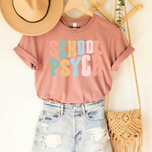 May include: A mauve t-shirt with the text "SCHOOL PSYCA" in colorful retro lettering. The shirt is hanging on a wooden rack with a straw bag and a brown hat.