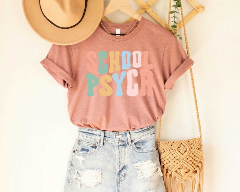 May include: A mauve t-shirt with the text "SCHOOL PSYCA" in colorful retro lettering. The shirt is hanging on a wooden rack with a straw bag and a brown hat.