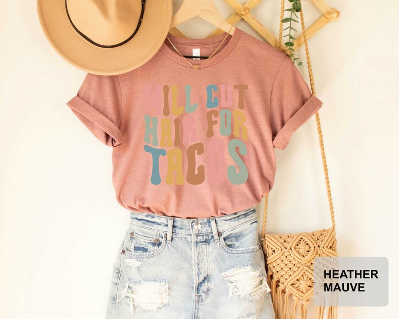 May include: A heather mauve t-shirt with the text "I'll cut hair for tacos" in colorful retro font.