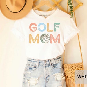 May include: White t-shirt with the text "GOLF MOM" in colorful letters with a golf ball graphic. The shirt is being modeled on a person wearing denim shorts.