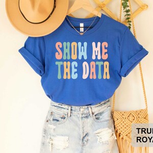 May include: A royal blue t-shirt with the text "Show Me The Data" in rainbow colors. The shirt is being modeled with a straw hat and denim shorts.