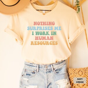 May include: A soft cream colored t-shirt with the text "Nothing Surprises Me I Work In Human Resources" in rainbow colors.