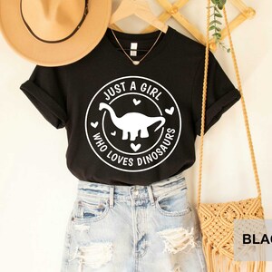 Puede incluir: Camiseta negra con un gr&aacute;fico circular blanco que dice "Just a girl who loves dinosaurs" con un dinosaurio blanco dentro del c&iacute;rculo.