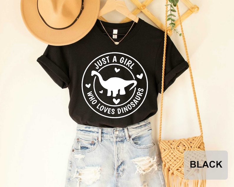 Puede incluir: Camiseta negra con un gr&aacute;fico circular blanco que dice "Just a girl who loves dinosaurs" con un dinosaurio blanco dentro del c&iacute;rculo.