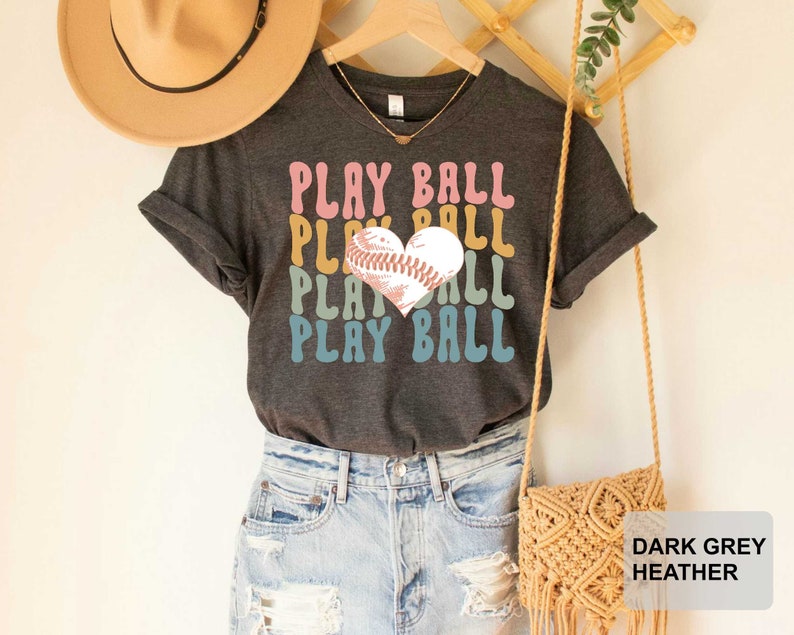 Peut inclure: Un t-shirt gris chin&eacute; fonc&eacute; avec un graphique d'un ballon de baseball en forme de c&oelig;ur et le texte "PLAY BALL" r&eacute;p&eacute;t&eacute; dans un sch&eacute;ma de couleurs arc-en-ciel.