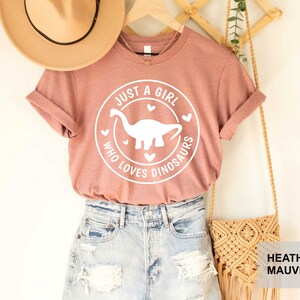 Puede incluir: Una camiseta de color malva claro con un gr&aacute;fico blanco que dice "Just a girl who loves dinosaurs" con una silueta de dinosaurio dentro de un c&iacute;rculo. La camiseta est&aacute; en una percha con un sombrero de paja y una bolsa tejida.
