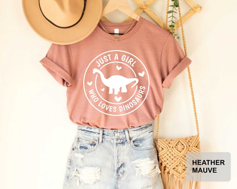 Puede incluir: Una camiseta de color malva claro con un gr&aacute;fico blanco que dice "Just a girl who loves dinosaurs" con una silueta de dinosaurio dentro de un c&iacute;rculo. La camiseta est&aacute; en una percha con un sombrero de paja y una bolsa tejida.