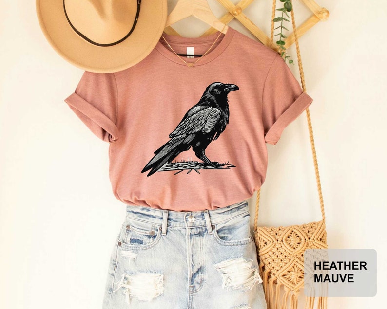 May include: A heather mauve t-shirt with a black and white illustration of a raven. The t-shirt is paired with light blue denim shorts and a tan crocheted bag.