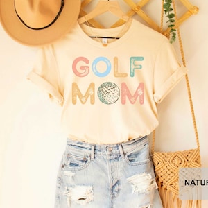 May include: A beige t-shirt with the text "GOLF MOM" in colorful letters and a golf ball graphic. The shirt is displayed on a mannequin with a straw hat and denim shorts.