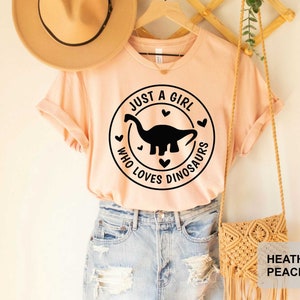 Puede incluir: Una camiseta de color melocot&oacute;n con un gr&aacute;fico negro de un dinosaurio y el texto "Just a girl who loves dinosaurs" en un c&iacute;rculo con corazones. La camiseta est&aacute; siendo modelada por una persona que lleva pantalones cortos de mezclilla y un sombrero de paja.