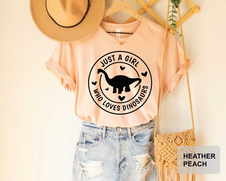 Puede incluir: Una camiseta de color melocot&oacute;n con un gr&aacute;fico negro de un dinosaurio y el texto "Just a girl who loves dinosaurs" en un c&iacute;rculo con corazones. La camiseta est&aacute; siendo modelada por una persona que lleva pantalones cortos de mezclilla y un sombrero de paja.