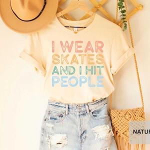 May include: A light beige t-shirt with the text "I wear skates and I hit people" in rainbow colours. The shirt is being modelled on a person wearing denim shorts and a brown hat.