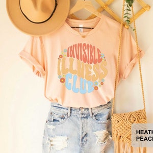 K&ouml;nnte beinhalten: Ein pfirsichfarbenes T-Shirt mit dem Text "Invisible Illness Club" in einer Retro-Schriftart mit bunten Blumen, die den Text umgeben. Das Shirt wird von einer Person in Jeans-Shorts getragen.