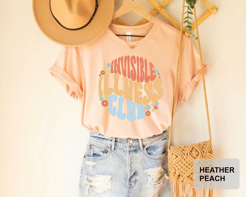 K&ouml;nnte beinhalten: Ein pfirsichfarbenes T-Shirt mit dem Text "Invisible Illness Club" in einer Retro-Schriftart mit bunten Blumen, die den Text umgeben. Das Shirt wird von einer Person in Jeans-Shorts getragen.