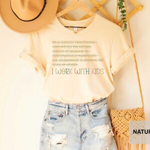 May include: A light beige t-shirt with the text "I'm a scientist practitioner who applies the natural science of behavior to systematically manipulate the environment to improve the lives of others. I work with kids." printed in colorful text.