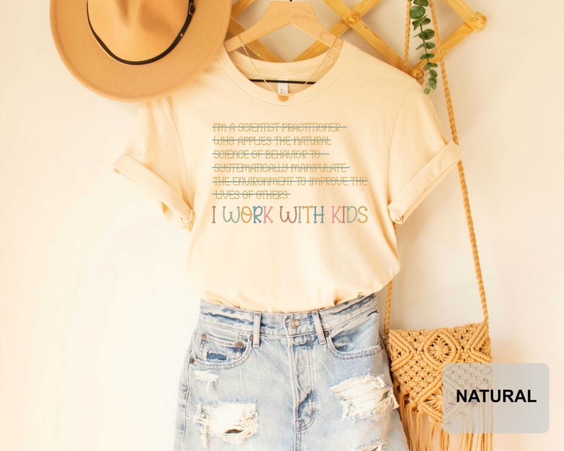 May include: A light beige t-shirt with the text "I'm a scientist practitioner who applies the natural science of behavior to systematically manipulate the environment to improve the lives of others. I work with kids." printed in colorful text.