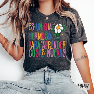 May include: Dark grey heather t-shirt with a colorful graphic of a flower with arms and legs. The text "Es un dia hermoso para aprender cosas nuevas" is printed on the shirt.