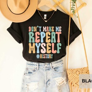May include: Black t-shirt with the text "Don't Make Me Repeat Myself #History" in colorful, retro-style lettering. The shirt is displayed on a mannequin with a straw hat and a woven bag.