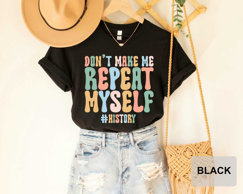 May include: Black t-shirt with the text "Don't Make Me Repeat Myself #History" in colorful, retro-style lettering. The shirt is displayed on a mannequin with a straw hat and a woven bag.