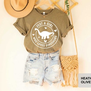 Puede incluir: Una camiseta verde con el texto "Just a girl who loves dinosaurs" y un gr&aacute;fico de dinosaurio blanco. La camiseta se combina con pantalones cortos de mezclilla y una bolsa de paja.