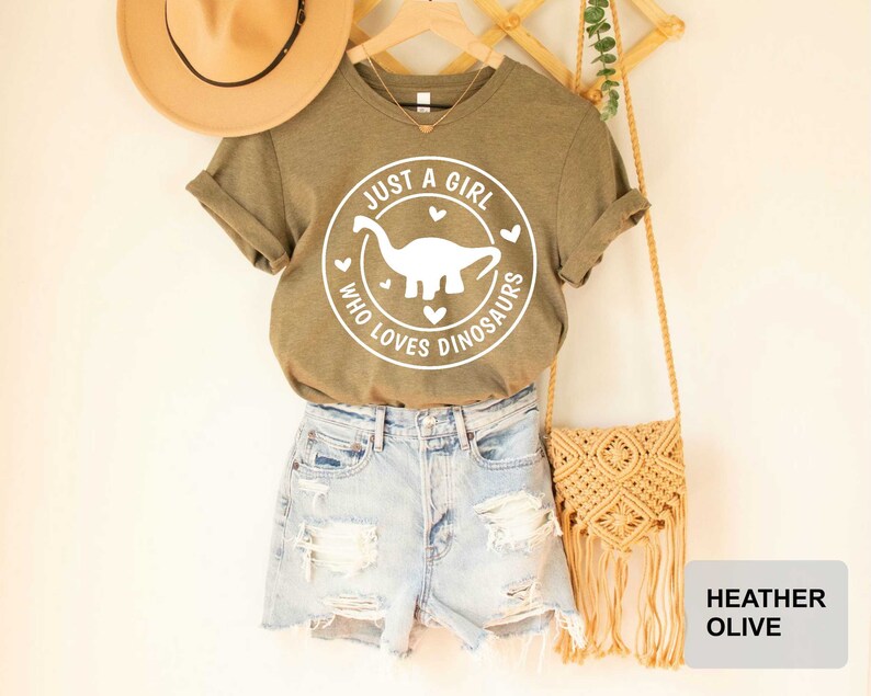 Puede incluir: Una camiseta verde con el texto "Just a girl who loves dinosaurs" y un gr&aacute;fico de dinosaurio blanco. La camiseta se combina con pantalones cortos de mezclilla y una bolsa de paja.