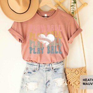 Peut inclure: Un t-shirt en mauve chin&eacute; avec un graphisme r&eacute;tro qui dit "Play Ball" avec un ballon de baseball en forme de c&oelig;ur.