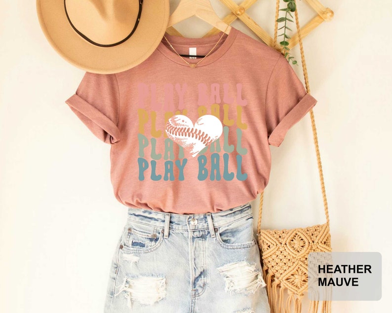 Peut inclure: Un t-shirt en mauve chin&eacute; avec un graphisme r&eacute;tro qui dit "Play Ball" avec un ballon de baseball en forme de c&oelig;ur.