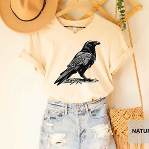 May include: A cream colored t-shirt with a black and white illustration of a raven on the front. The shirt is being modeled on a person wearing blue denim shorts and a brown hat.