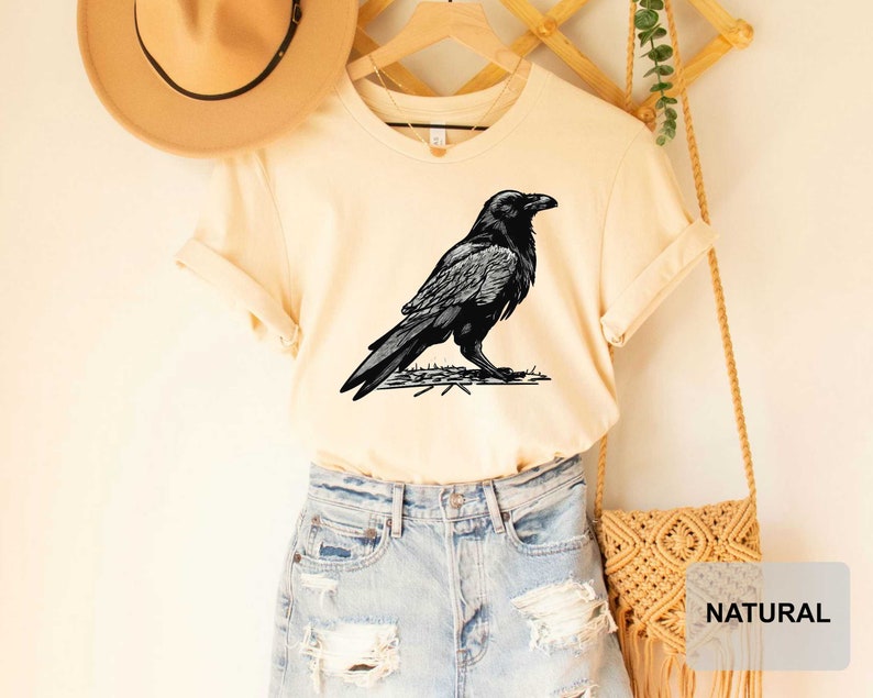 May include: A cream colored t-shirt with a black and white illustration of a raven on the front. The shirt is being modeled on a person wearing blue denim shorts and a brown hat.