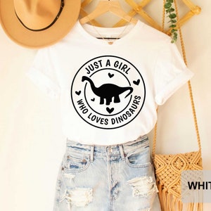 Puede incluir: Camiseta blanca con un gr&aacute;fico circular negro que dice "Just a girl who loves dinosaurs" con una silueta de dinosaurio dentro del c&iacute;rculo.