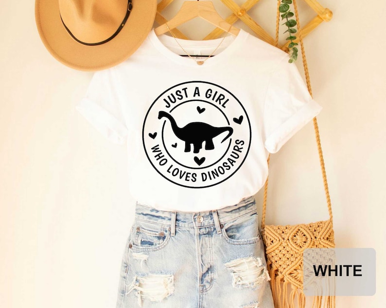 Puede incluir: Camiseta blanca con un gr&aacute;fico circular negro que dice "Just a girl who loves dinosaurs" con una silueta de dinosaurio dentro del c&iacute;rculo.