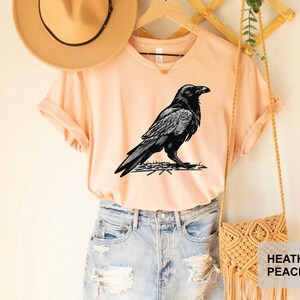 May include: A heather peach t-shirt with a black and white illustration of a raven on a branch. The shirt is being modeled on a person wearing denim shorts.