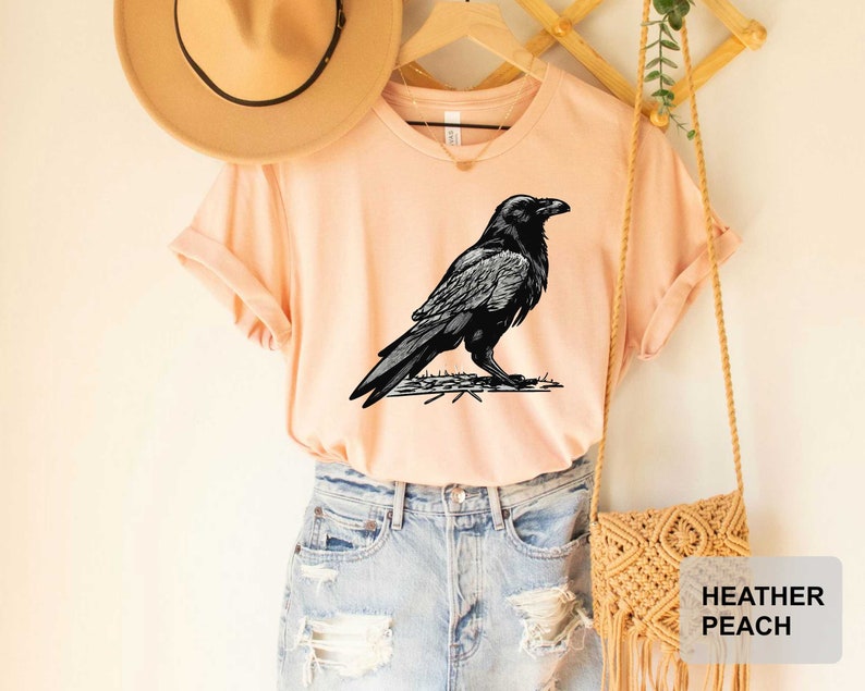 May include: A heather peach t-shirt with a black and white illustration of a raven on a branch. The shirt is being modeled on a person wearing denim shorts.