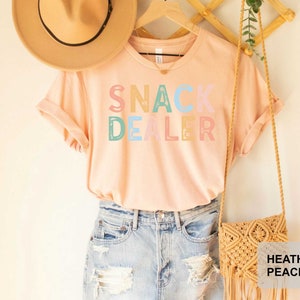 Puede incluir: Una camiseta rosa claro con el texto "SNACK DEALER" en letras de colores. La camiseta se est&aacute; modelando con un sombrero de paja y un bolso de crochet.