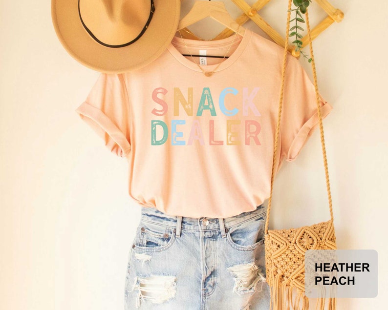 Puede incluir: Una camiseta rosa claro con el texto "SNACK DEALER" en letras de colores. La camiseta se est&aacute; modelando con un sombrero de paja y un bolso de crochet.