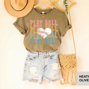 Peut inclure: Un t-shirt en couleur olive chin&eacute; avec le texte "PLAY BALL" en couleurs arc-en-ciel et un ballon de baseball en forme de c&oelig;ur. Le t-shirt est port&eacute; par une personne portant un short en jean.