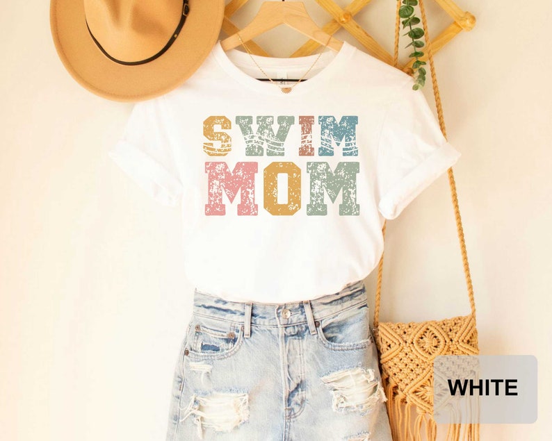 Pu&ograve; includere: Maglietta bianca con il testo "Swim Mom" in lettere colorate e consumate. La maglietta &egrave; indossata sopra un paio di pantaloncini di jeans blu chiaro.