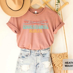 May include: A heather mauve t-shirt with the text "Teacher? Educational Ninja" in a retro font. The shirt is paired with denim shorts and a woven bag.