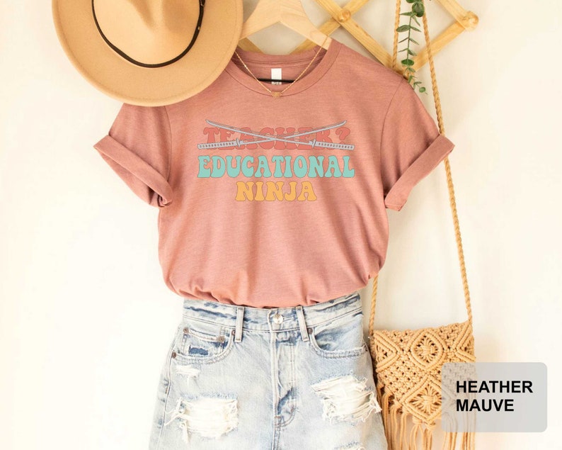 May include: A heather mauve t-shirt with the text "Teacher? Educational Ninja" in a retro font. The shirt is paired with denim shorts and a woven bag.