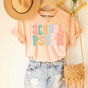 May include: A light pink t-shirt with the words "SCHOOL PSYCH" in colorful, retro-style lettering.